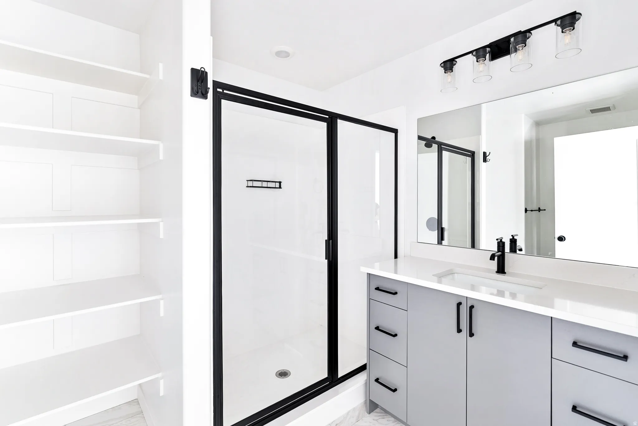 Built-in shelving, glass-enclosed shower with black trim, vanity with light gray cabinetry and black hardware, white countertop, and a large wall-mounted mirror