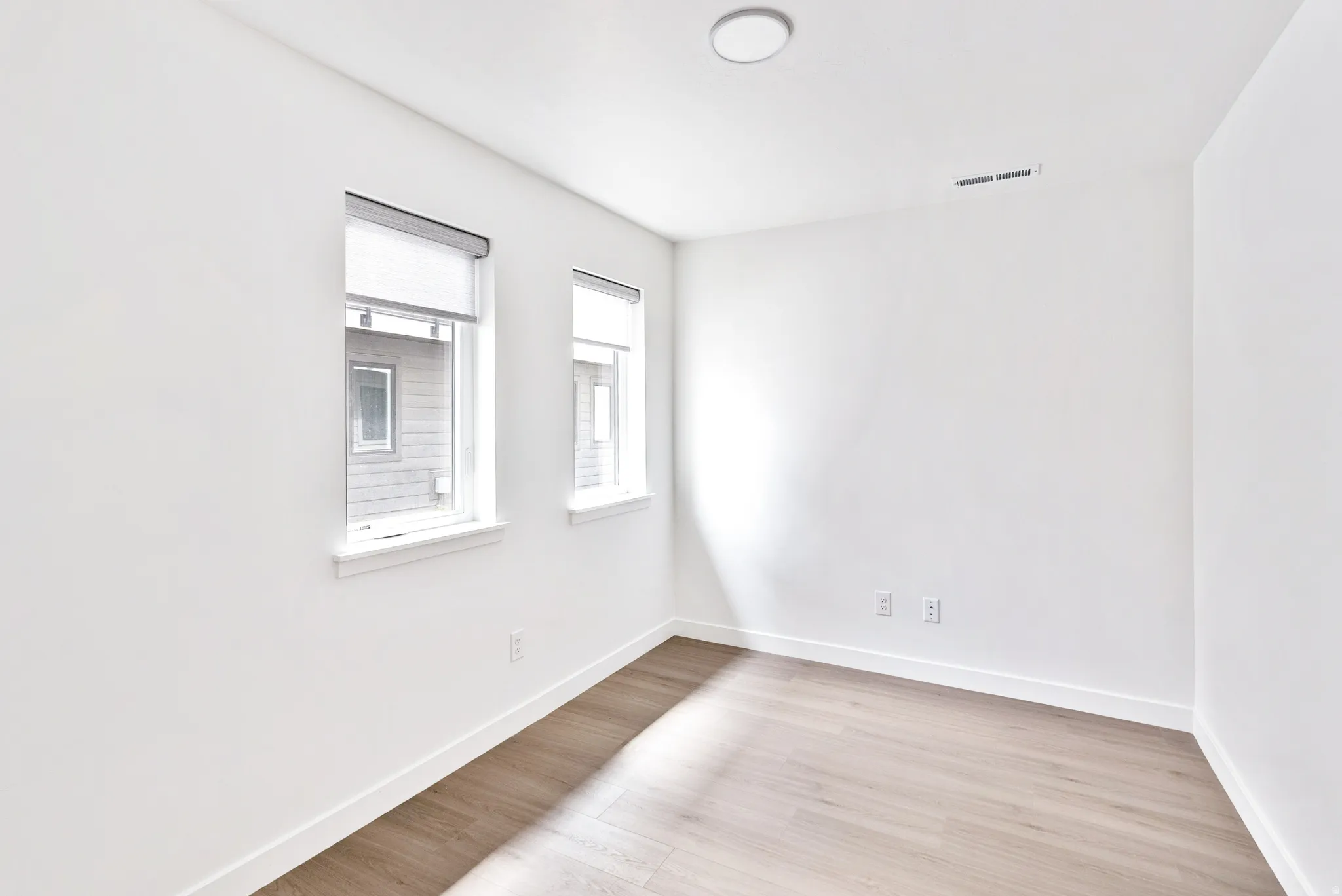 Light wood-finish flooring, white walls, two windows with roller shades, and a flush-mount ceiling light