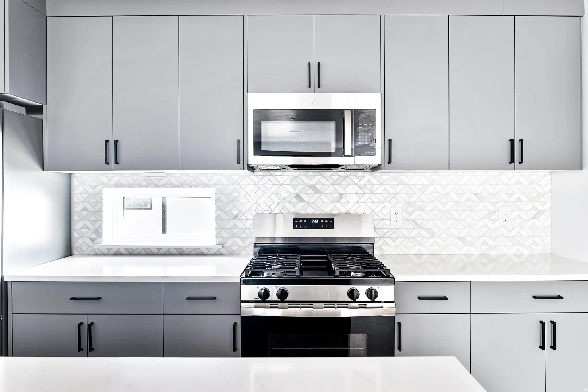 Contemporary kitchen featuring light gray cabinetry, stainless steel appliances, a unique patterned backsplash, and white countertops