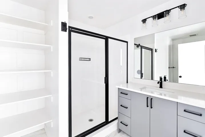 Built-in shelving, matte black framed glass shower, light gray vanity with black hardware, white rectangular undermount sink, and a four-light vanity fixture