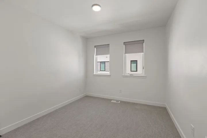 Recessed ceiling light, two windows with roller shades, light grey carpeting, and white baseboards