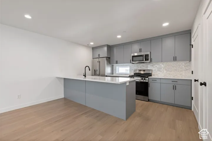 Kitchen featuring gray cabinetry, stainless steel appliances, a white countertop island, a geometric tile backsplash, and wood-finish flooring