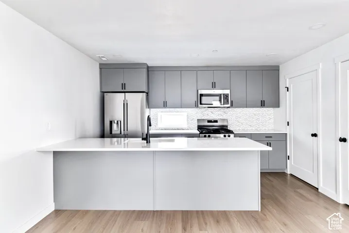 Kitchen featuring light wood-finish flooring, a white countertop island, stainless steel appliances, gray cabinetry, and a patterned tile backsplash