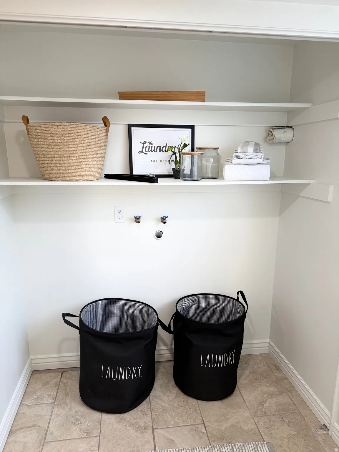 Laundry closet with two built-in shelves, wall-mounted electrical outlet, and tiled flooring