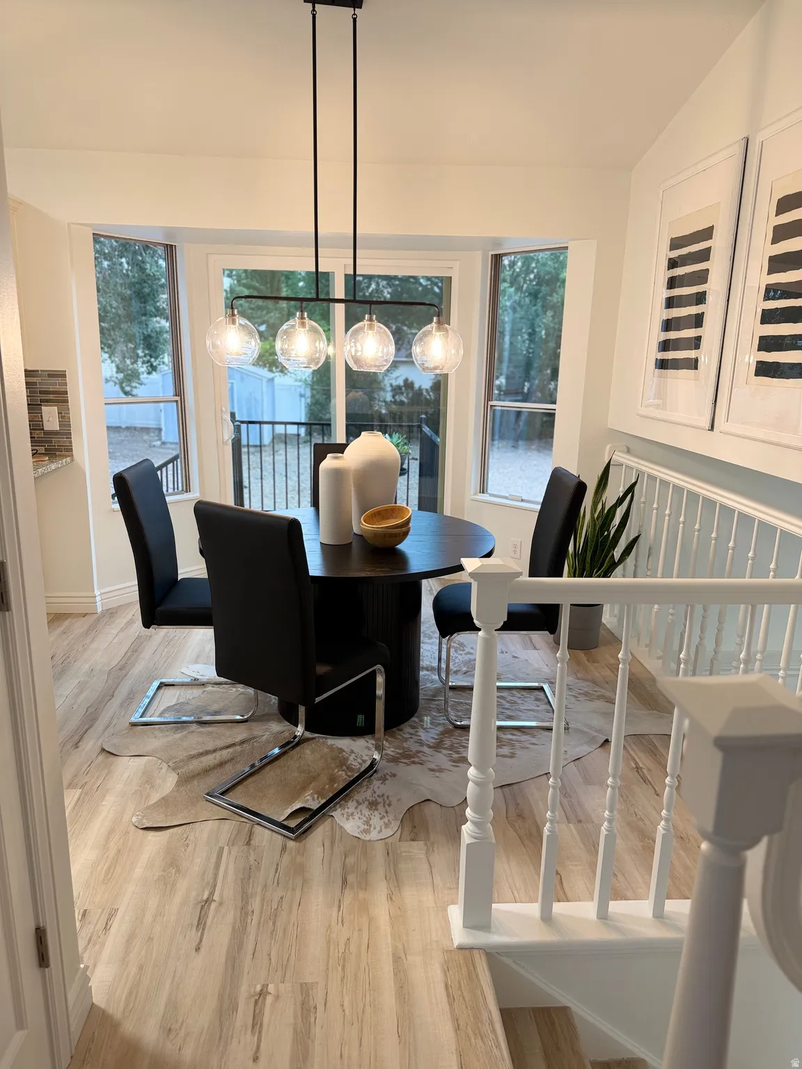 Recessed bay window, modern linear chandelier, light wood-finish flooring, white banister with turned spindles, and a tiled accent wall