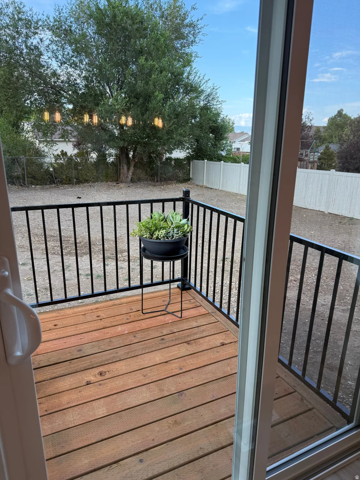 Wood-plank deck with black metal railing, featuring a sliding glass door and a spacious backyard with a white privacy fence and mature trees