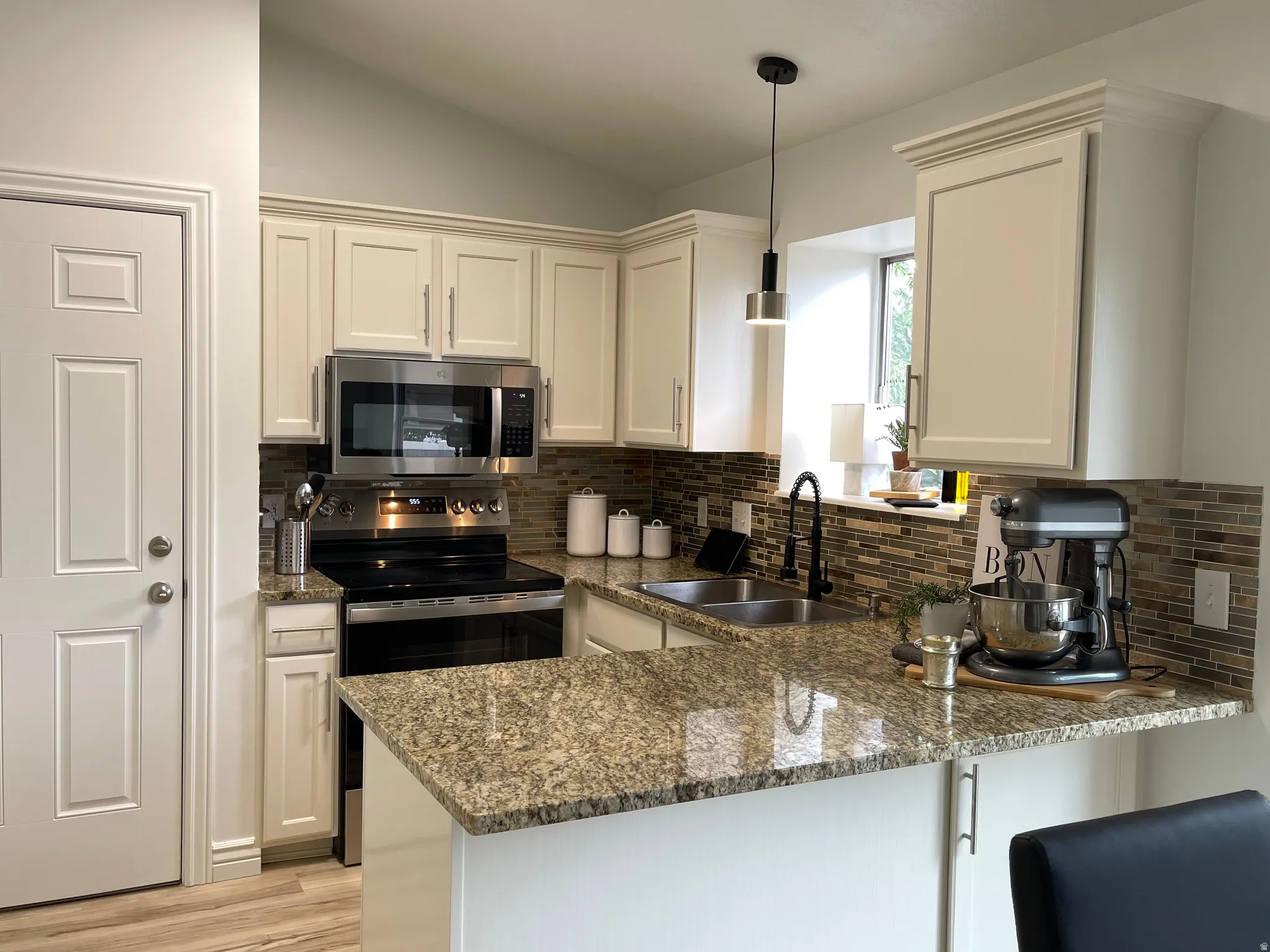 Kitchen featuring light cabinetry, stainless steel appliances, a double basin sink, mosaic tile backsplash, and a peninsula with a stone countertop