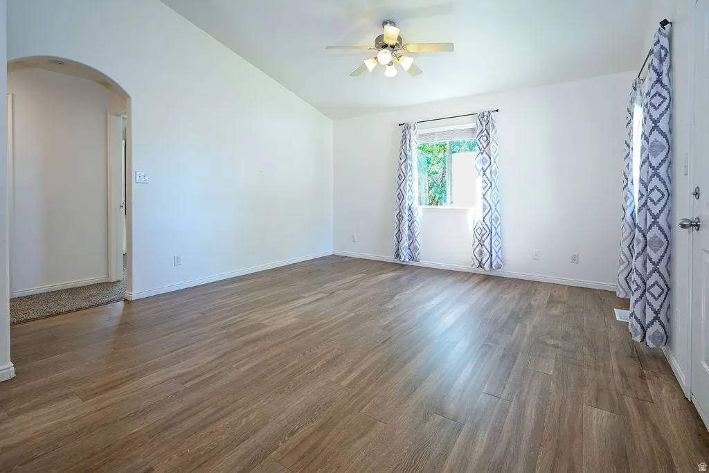 Vaulted ceiling, wood-finish flooring, ceiling fan with light fixture, arched doorway, and a window with trim