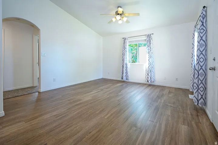 Vaulted ceiling, wood-finish flooring, ceiling fan with light fixture, arched doorway, and a window with trim
