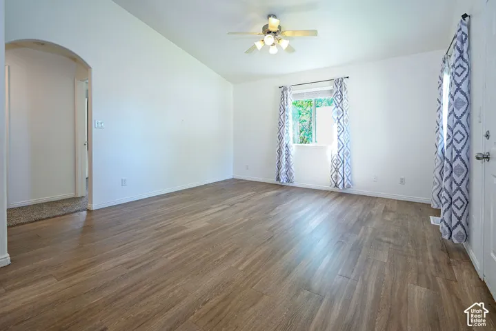 Vaulted ceiling, wood-finish flooring, ceiling fan with light fixture, arched doorway, and a window with trim