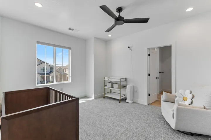 Bright nursery with abundant natural light from large window, gray carpet flooring, ceiling fan with remote control, dark wood crib, open shelving unit, and direct access to adjacent bathroom with white vanity and vessel sink