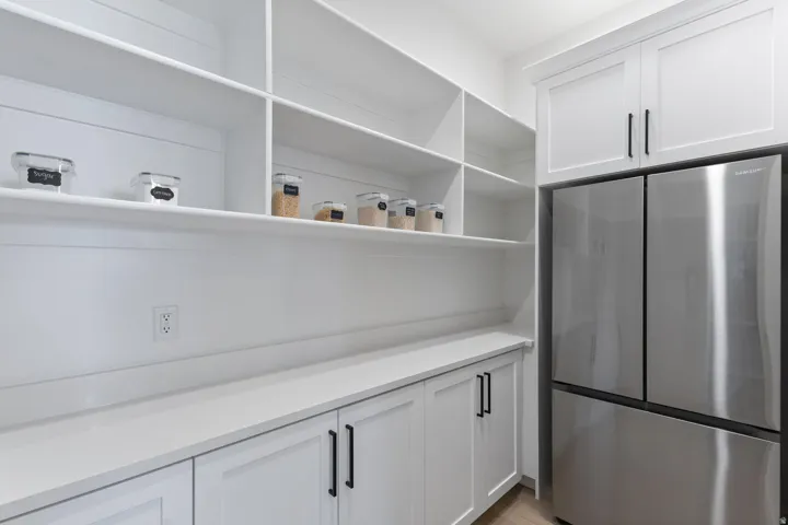 Modern pantry featuring extensive open shelving with integrated storage, white cabinetry with black hardware, stainless steel refrigerator with French-door configuration, quartz countertop, and recessed lighting throughout