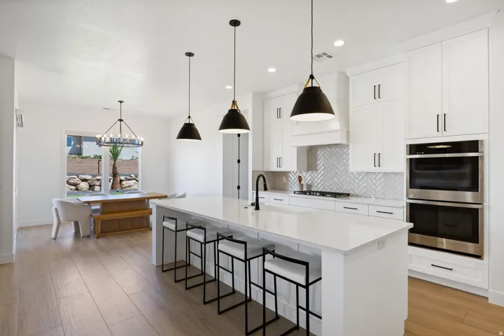 White cabinetry with matte black hardware, integrated double ovens, herringbone tile backsplash, large central island, and wood-finish flooring