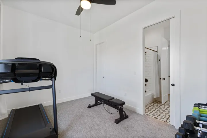 White interior walls, ceiling fan with integrated lighting, built-in closet with white door, gray carpeted flooring, and an adjoining bathroom featuring a white subway tile shower and patterned floor tile