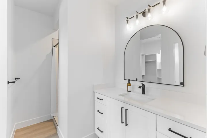 Modern primary bathroom featuring a floating vanity with dual sinks, white cabinetry with matte black hardware, quartz countertop, and an arched mirror with integrated lighting