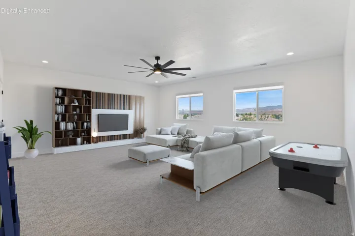 Spacious recreation room featuring neutral carpet flooring, a ceiling fan with integrated lighting, built-in entertainment cabinetry with shelving and media display, large windows with panoramic outlooks, and a game table area