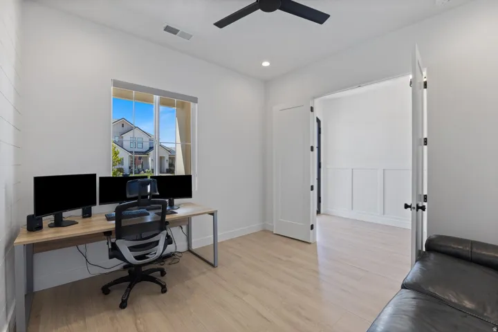 Bright home office featuring expansive windows with natural light, wood-finish flooring throughout, dual monitor setup capability with ample desk space, ceiling fan, and direct access to a spa-style ensuite bathroom with glass-enclosed shower and black ha