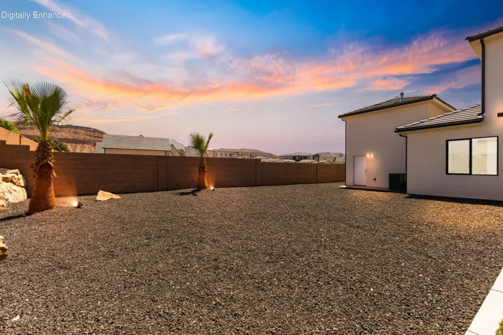 Expansive rear yard featuring dark gravel, privacy wall, mature palm trees, and exterior lighting