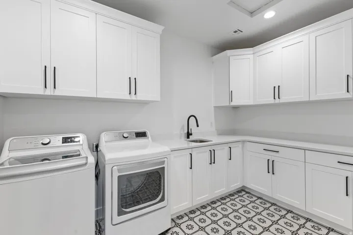 Modern laundry room featuring white shaker-style cabinetry with matte black hardware, a stainless steel sink with matte black faucet, geometric patterned tile flooring, and space for full-size front-load washer and dryer appliances