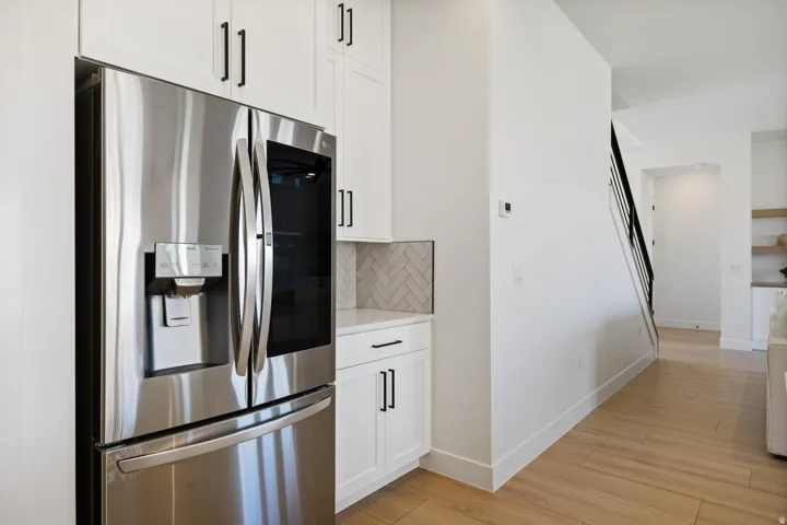 Stainless steel refrigerator, white shaker cabinetry, herringbone tile backsplash, wood-finish flooring, and a black wrought iron staircase railing
