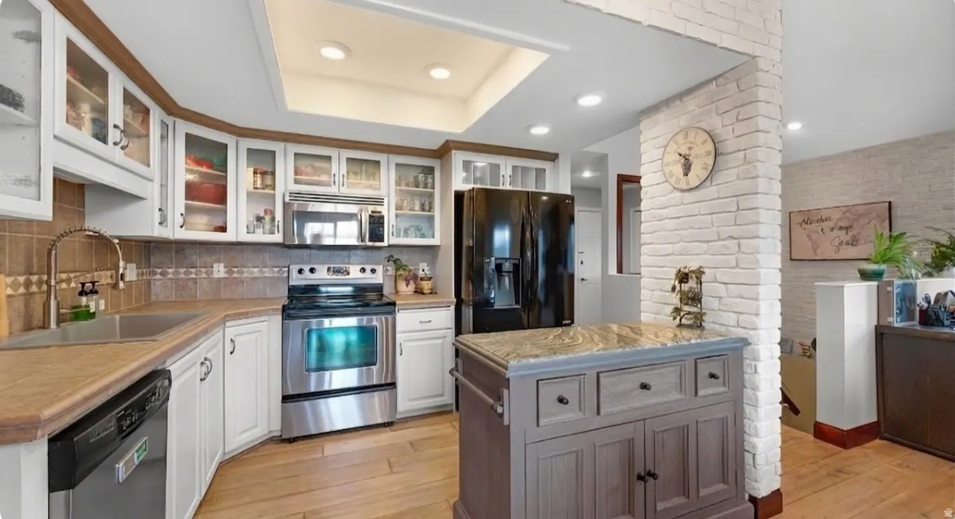 Kitchen with stainless steel appliances, wood-finish flooring, white cabinetry, a stone-top island, and a brick accent wall