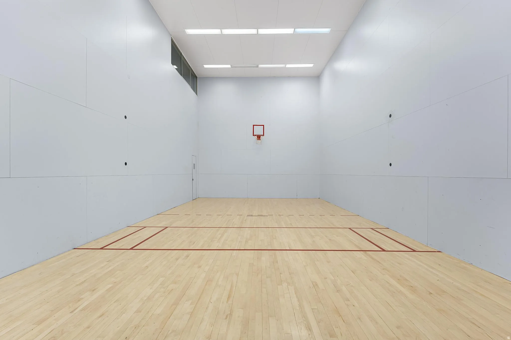 Indoor court featuring light wood flooring, white panel walls, a basketball hoop, overhead fluorescent lighting, and a high-set window