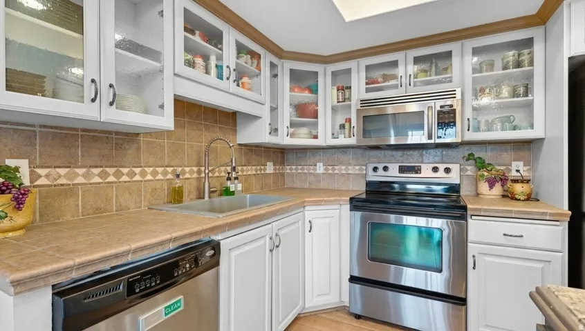 White cabinetry with glass-front upper doors, stainless steel appliances, tile backsplash with decorative accent, and a single basin stainless steel sink with a gooseneck faucet