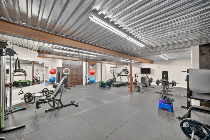 Expansive fitness space featuring exposed metal ceiling, recessed linear lighting, rubber flooring, and large wall-mounted mirrors