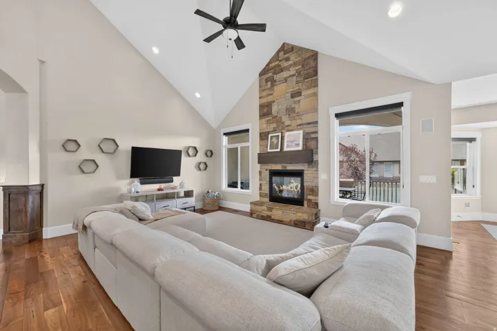 Vaulted ceilings with a ceiling fan, stone-clad fireplace with a wood mantle, recessed lighting, large windows, and wood-finish flooring