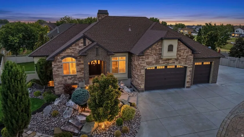 Stone and stucco exterior with dark brown garage doors, a large concrete driveway, and a tiered rock garden with landscaping