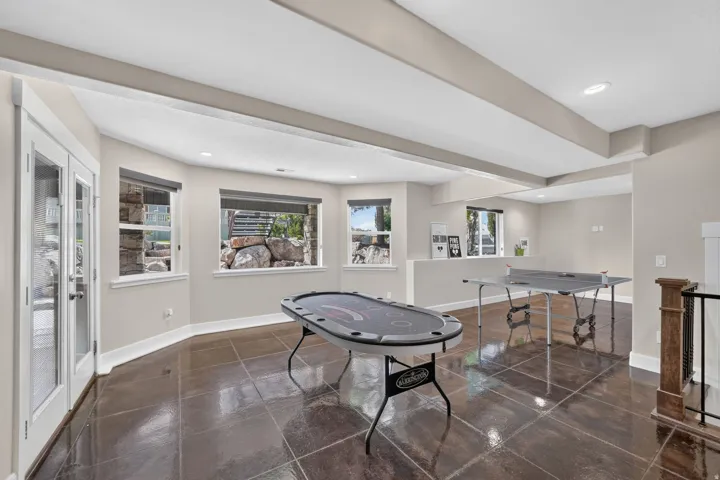 Spacious lower-level area with large format tile flooring, multiple windows providing natural illumination, a built-in half-wall, and exposed ceiling beams