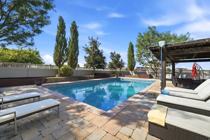 Swimming pool with stone coping and surrounding paver patio, featuring a dark wood pergola
