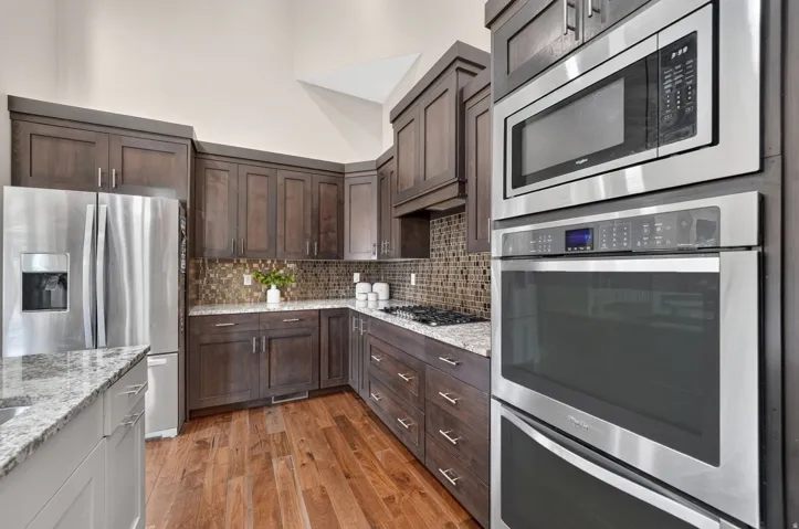 Kitchen featuring wood-finish flooring, dark wood cabinetry, light granite countertops, stainless steel appliances, and a mosaic tile backsplash