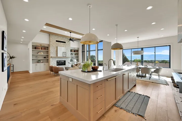 Expansive open-concept living area featuring wood-finish flooring, a contemporary kitchen island with a light-toned countertop, a stone-clad fireplace, built-in shelving, and large windows