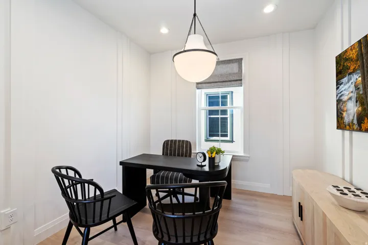 Light wood-finish flooring, white walls with vertical paneling, a large window with a Roman shade, and a pendant light fixture