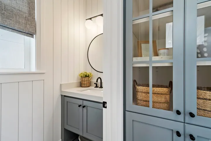 Board and batten wall paneling, vanity with a white countertop and undermount sink, matte black faucet, round wall-mounted mirror, and glass-front cabinetry
