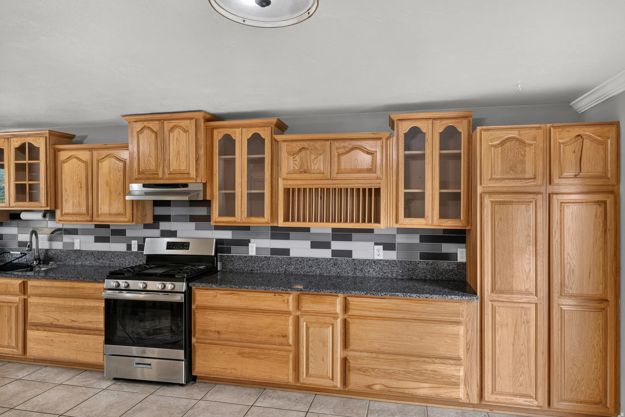 Wood-finish cabinetry, dark speckled countertops, stainless steel range with overhead hood, and a tiled backsplash