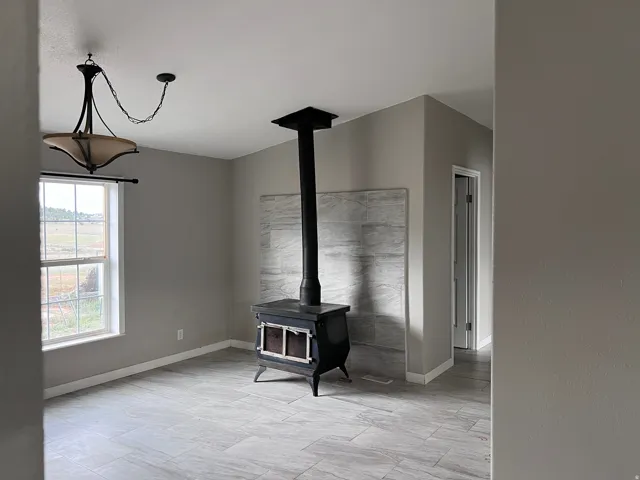 Wood-burning stove with a tiled backdrop, large window, ceiling-mounted light fixture, and tile flooring