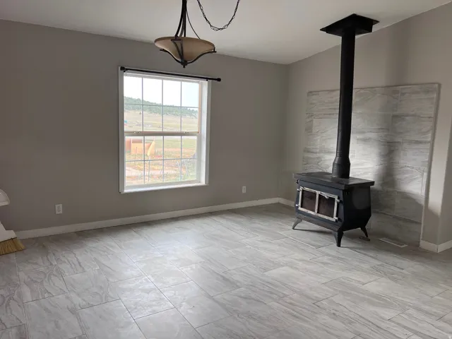Wood-burning stove with a tiled backdrop, large window, overhead light fixture, and tile flooring