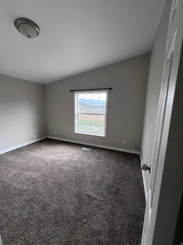 Neutral tone wall paint, vaulted ceiling, window with dark curtain rod, brown carpeting, and white trim