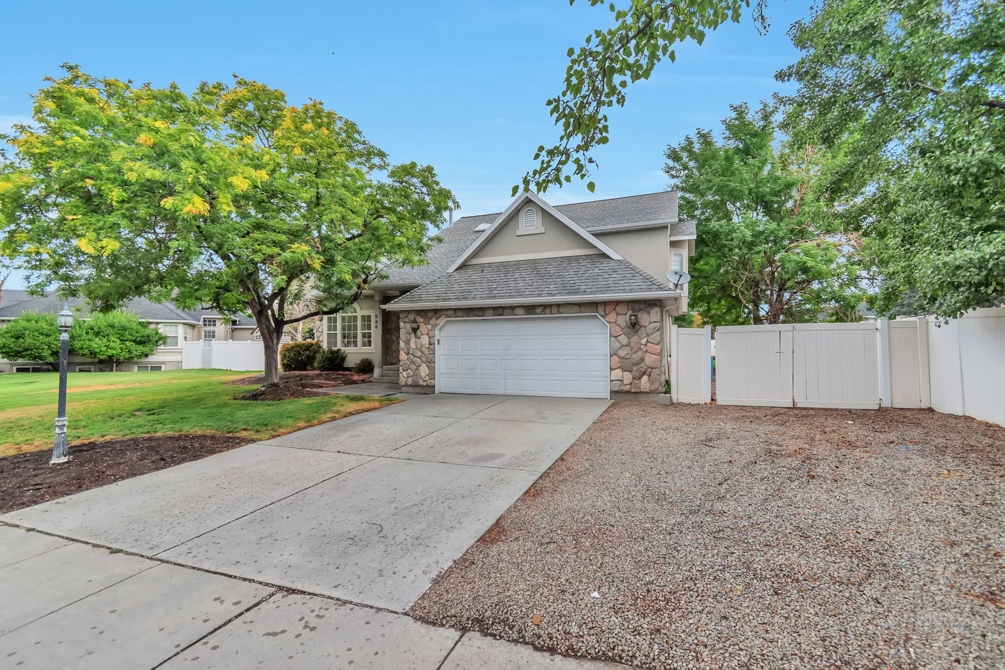 Stone-clad facade with an attached two-car garage, mature trees, concrete driveway, and a fenced side yard