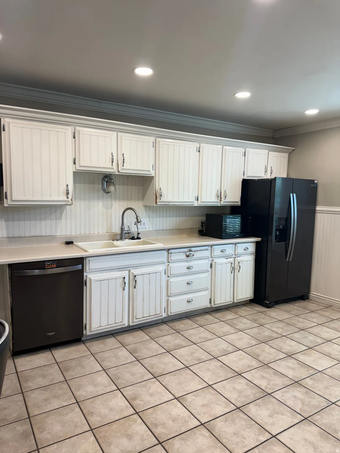 Kitchen featuring white cabinetry with beadboard detailing, light-toned countertops, a double basin sink, and a black dishwasher