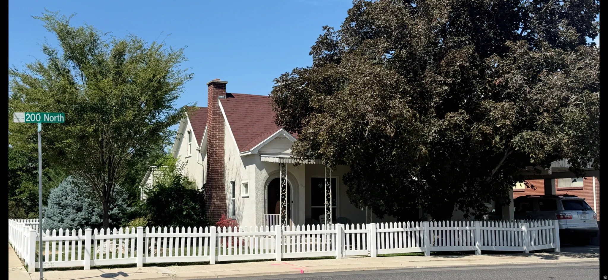 Charming single-family residence featuring a white picket fence, mature landscaping, a prominent brick chimney, an arched porch entry, and a dark shingle roof