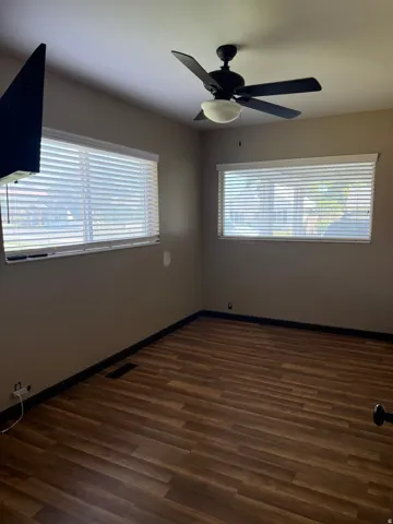 Spacious room featuring wood-finish flooring, two large windows with blinds, a black ceiling fan with an integrated light fixture, and neutral-toned walls with dark trim