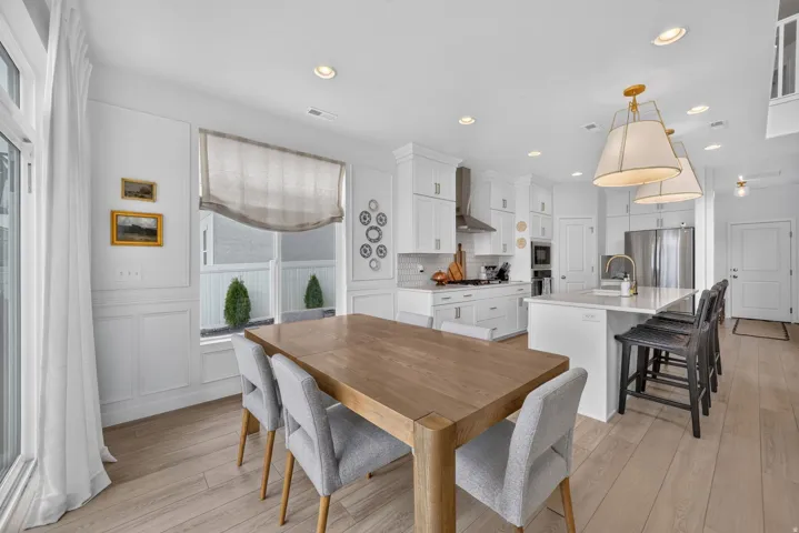 Open-concept living area with light wood-finish flooring, white paneled walls, a white kitchen island with seating, white shaker-style cabinetry, and stainless steel appliances