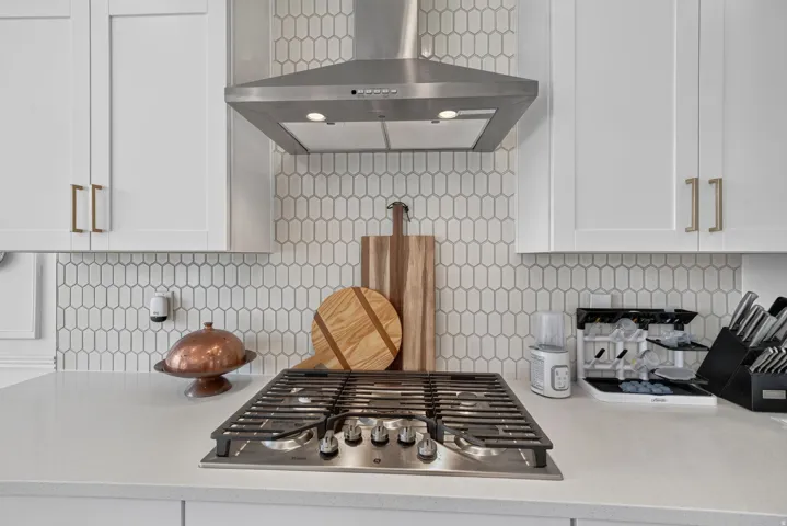 White shaker cabinetry with gold-finish hardware, stainless steel range hood, picket tile backsplash, built-in gas cooktop, and light-tone countertop