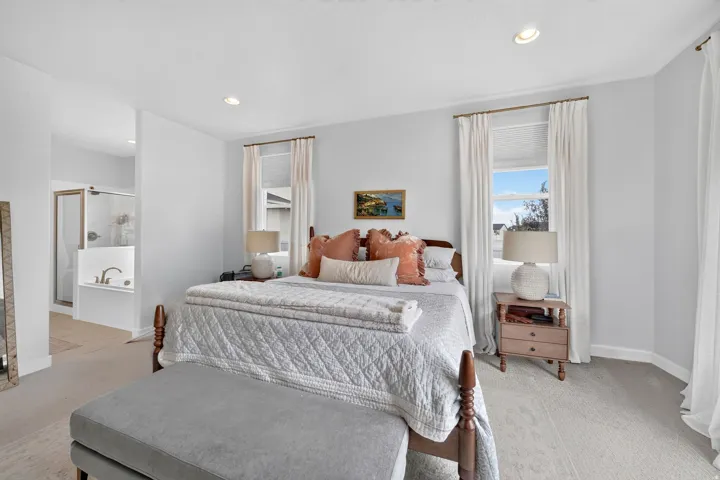 Recessed lighting, light-toned carpeting, two windows with white window treatments, and a built-in soaking tub with a glass-enclosed shower