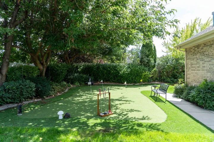 Putting green with multiple holes, brick exterior, mature trees, and a concrete pathway