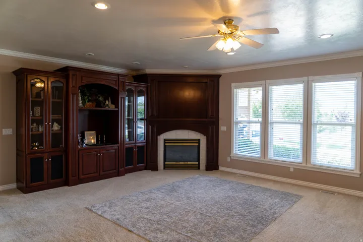 Built-in cabinetry with glass-front display, fireplace with white mantle, three large windows, recessed lighting, and a ceiling fan