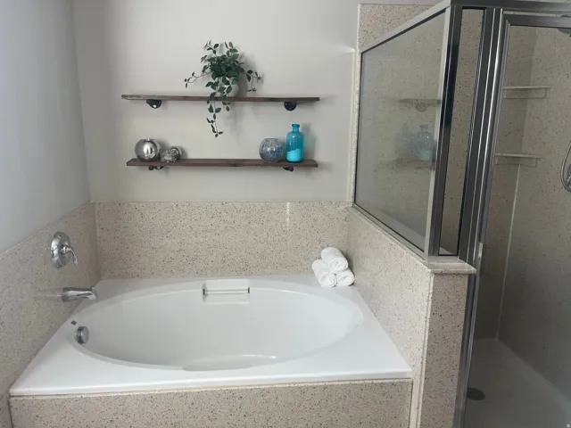 Built-in soaking tub with chrome fixtures, enclosed shower with glass surround, and two wall-mounted wood shelves